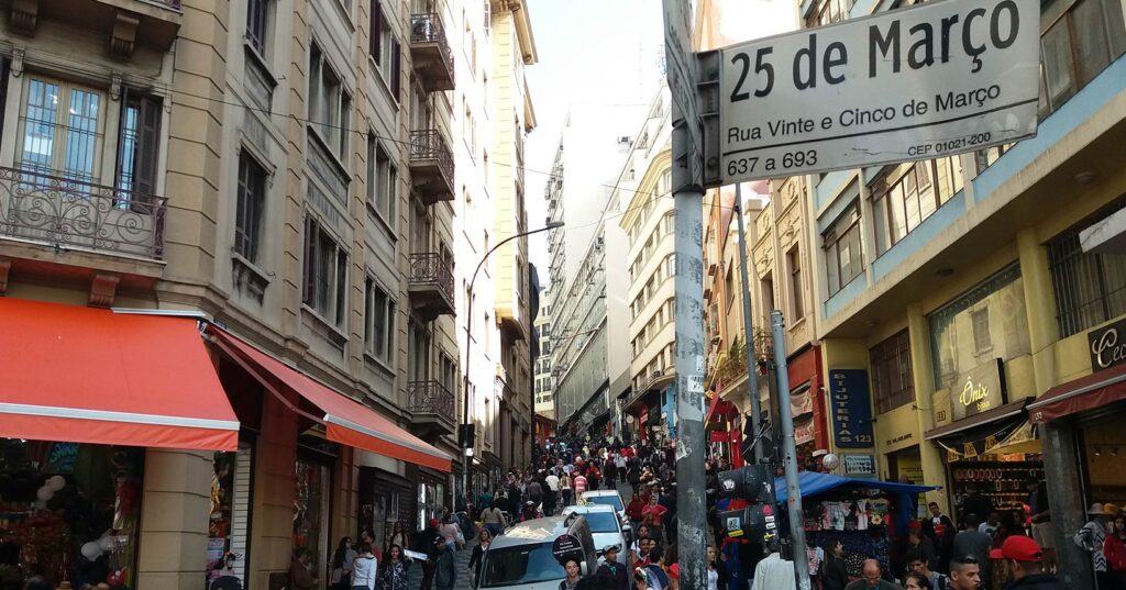 Movimentação na Rua 25 de Março em São Paulo, com várias lojas e consumidores em busca de compras.